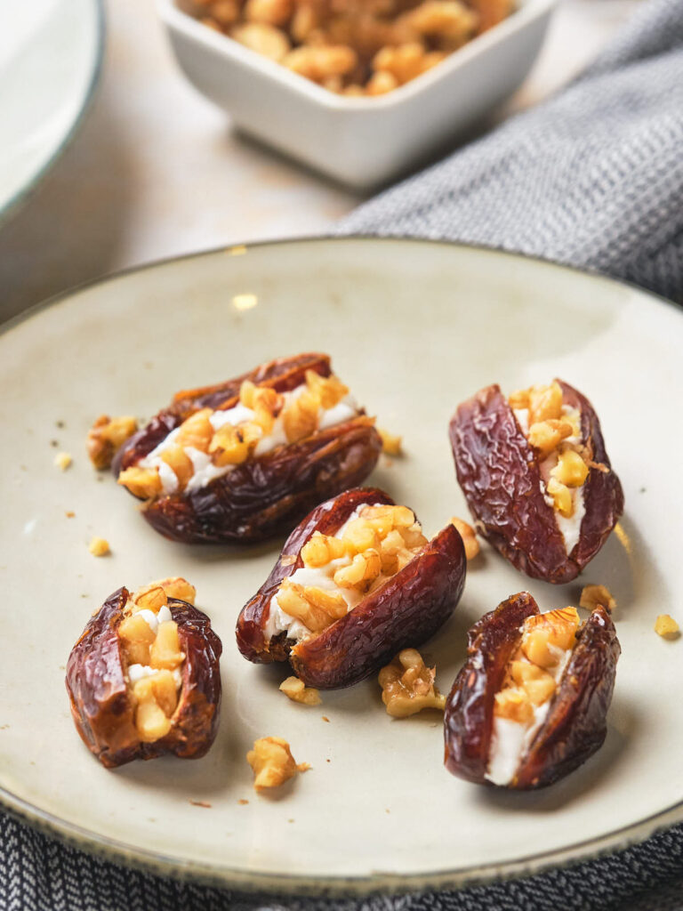 Five dates on a plate, stuffed with walnuts, with more walnuts in a dish in the background.