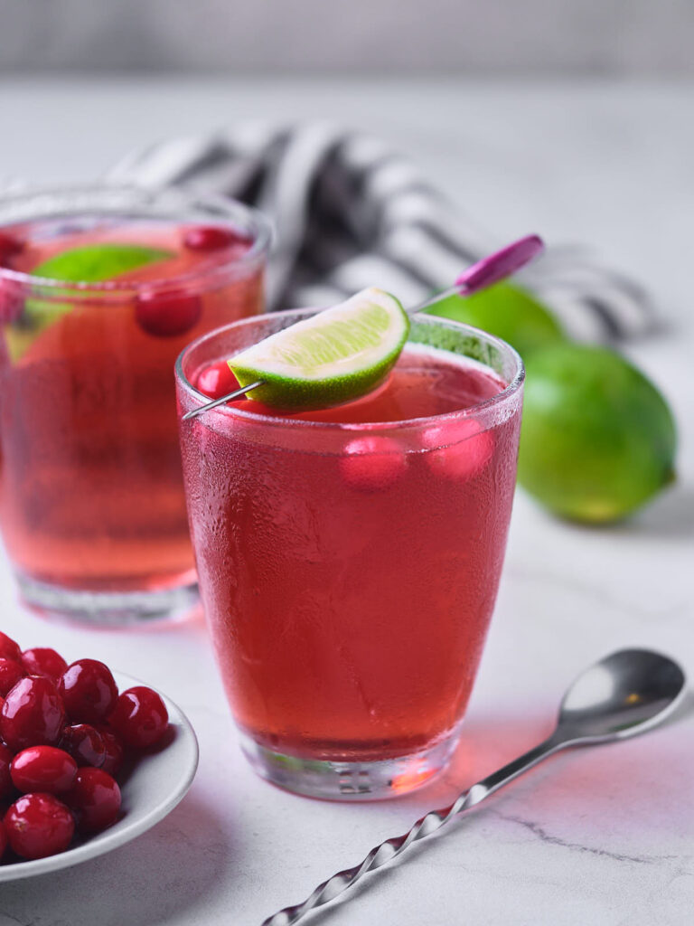 Two glasses of vodka cranberry cranberries and lime wedges on a marble surface.