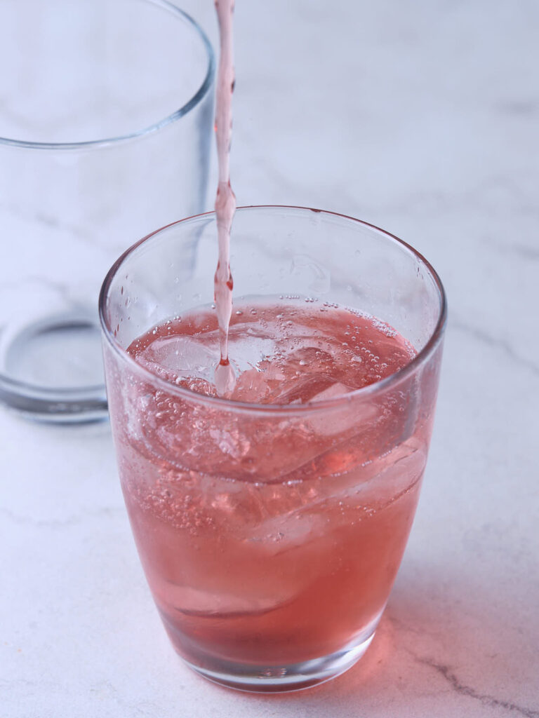 A pink drink is being poured into a clear glass with ice cubes.