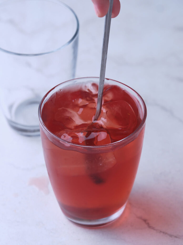 A hand stirring a red iced drink in a glass with a metal spoon.