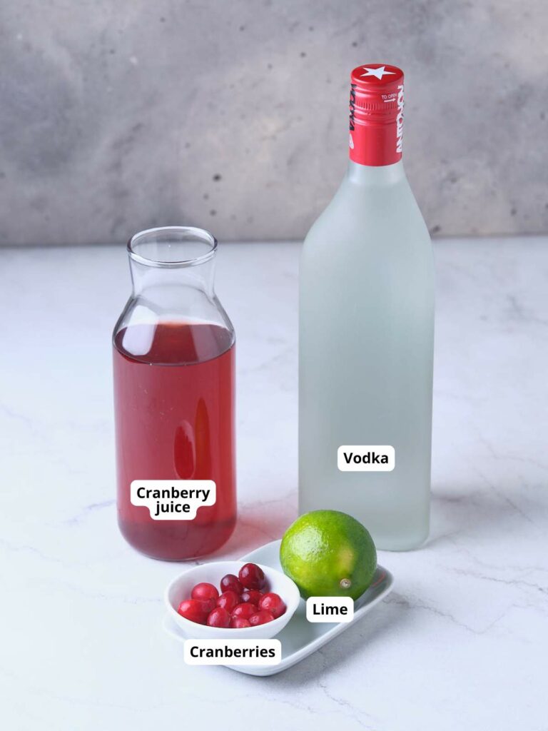 A bottle of vodka, a small jug of cranberry juice, and a plate with lime and cranberries are arranged on a marble surface.