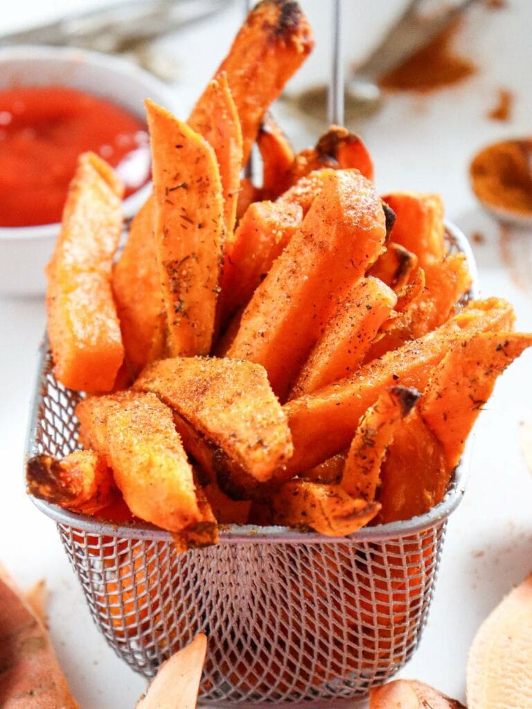 A basket of crispy seasoned sweet potato fries is placed on a table, with a small bowl of dipping sauce in the background.