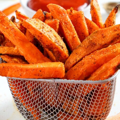 A metal basket filled with seasoned sweet potato fries sits in front of a bowl of red dipping sauce.