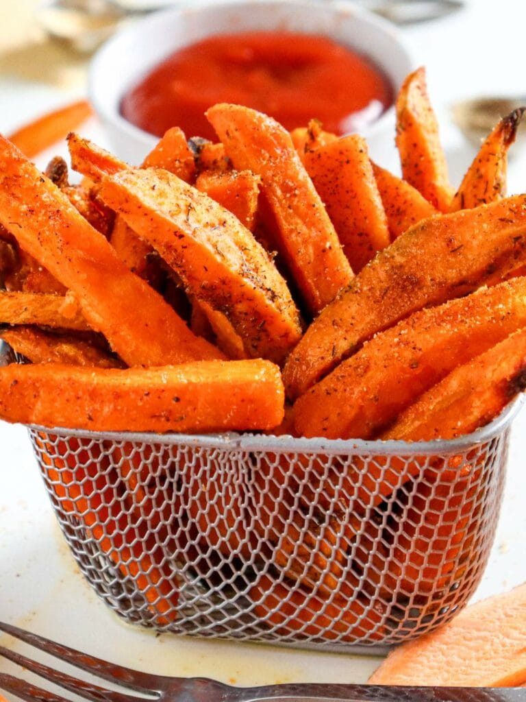 A metal basket filled with seasoned sweet potato fries sits in front of a bowl of red dipping sauce.