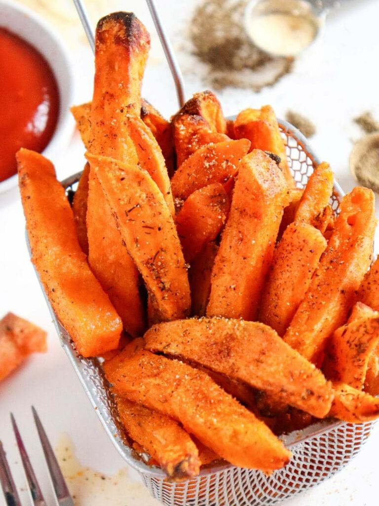 A basket of seasoned sweet potato fries on a table, with a fork and a bowl of ketchup in the background.