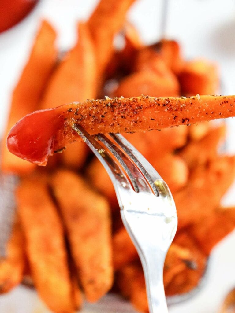 Close-up of a fork holding a seasoned sweet potato fry dipped in ketchup, with a basket of fries blurred in the background.