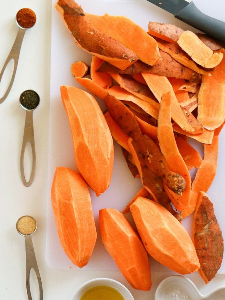 Peeled sweet potatoes and their skins on a cutting board beside measuring spoons containing various spices.