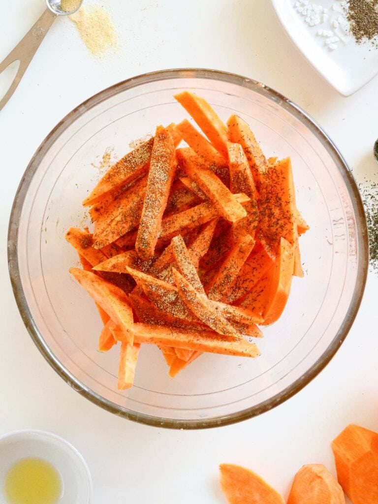 Cut sweet potato fries seasoned with spices in a glass bowl, surrounded by small dishes with oil and salt on a white surface.