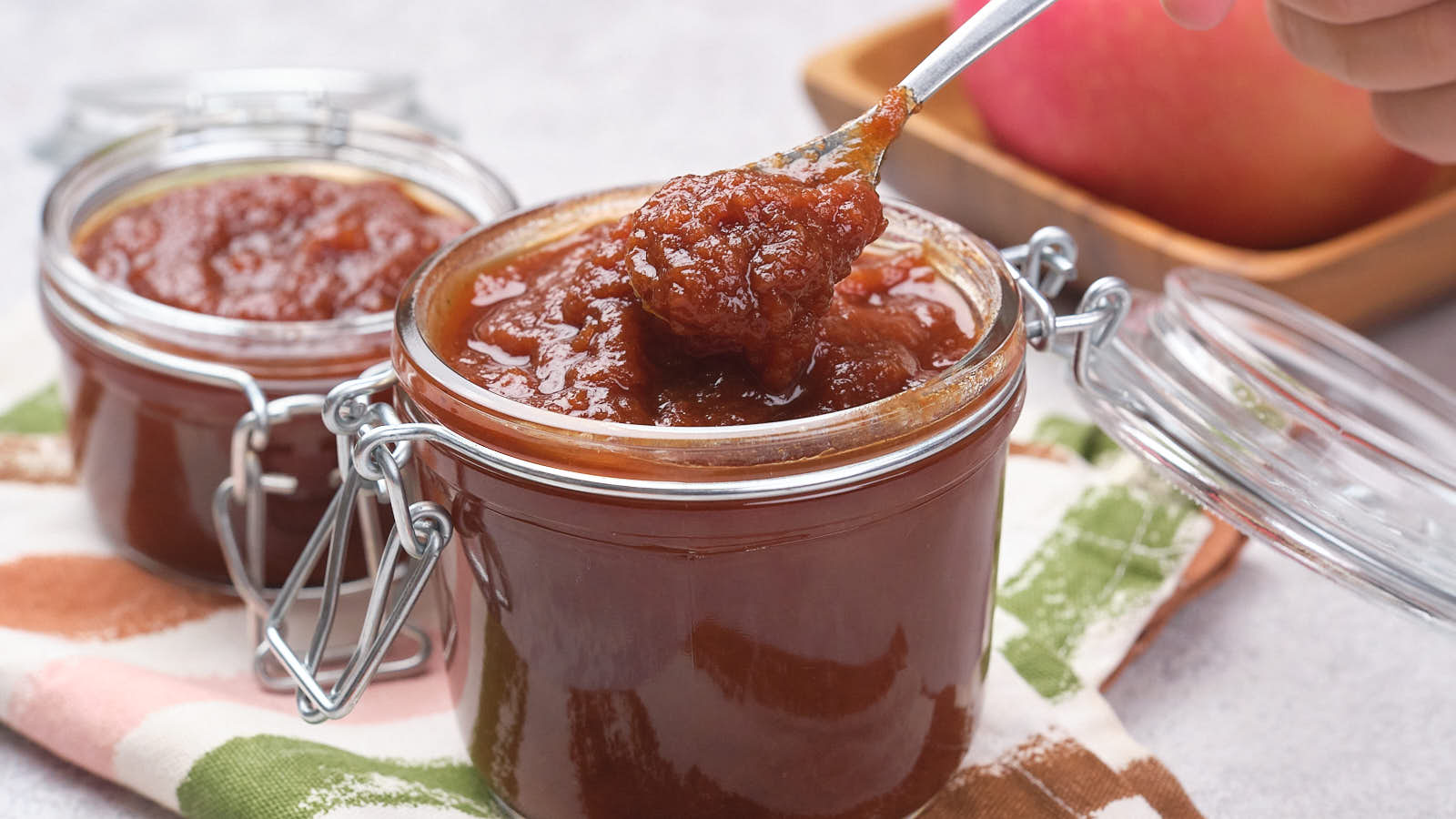 Two glass jars of apple butter, one open with a spoonful being lifted, placed on a patterned cloth.