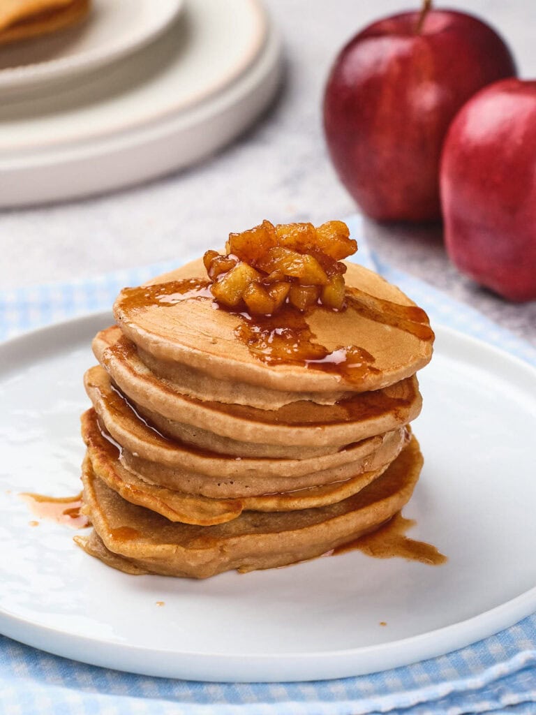 A stack of pancakes topped with caramelized apples and syrup is on a white plate.