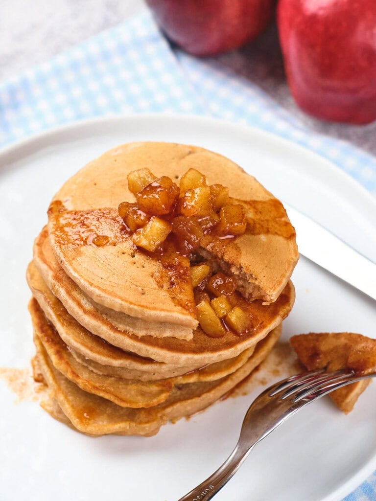 A stack of pancakes topped with caramelized apples on a white plate. A fork and knife sit beside the pancakes, and apples are visible in the background.