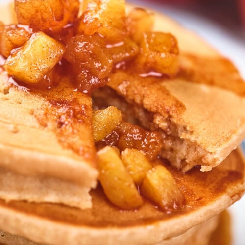 A stack of pancakes topped with caramelized apple pieces, with a slice cut out, revealing more apple filling inside.