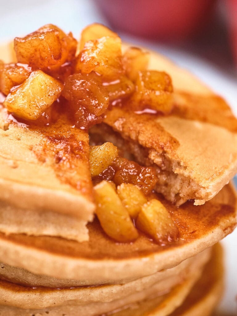 A stack of pancakes topped with caramelized apple pieces, with a slice cut out, revealing more apple filling inside.