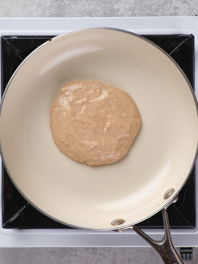 A pancake batter is spreading on a non-stick frying pan placed on a stove.