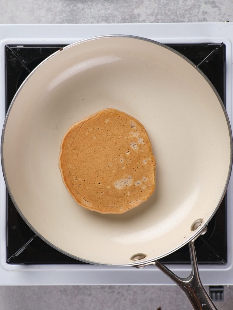 A single pancake cooking in a white frying pan on a stovetop.