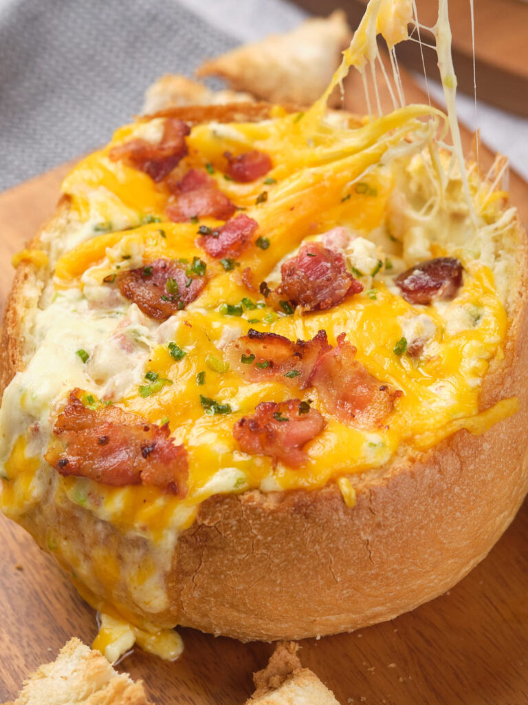 Cheesy dip in a bread bowl topped with bacon and herbs, with melted cheese stretching as a piece is lifted.