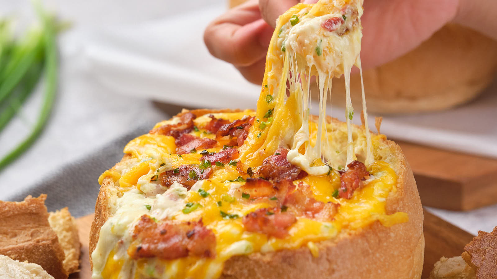 A person pulls a cheesy topping from a bread bowl filled with melted cheese and bacon bits.