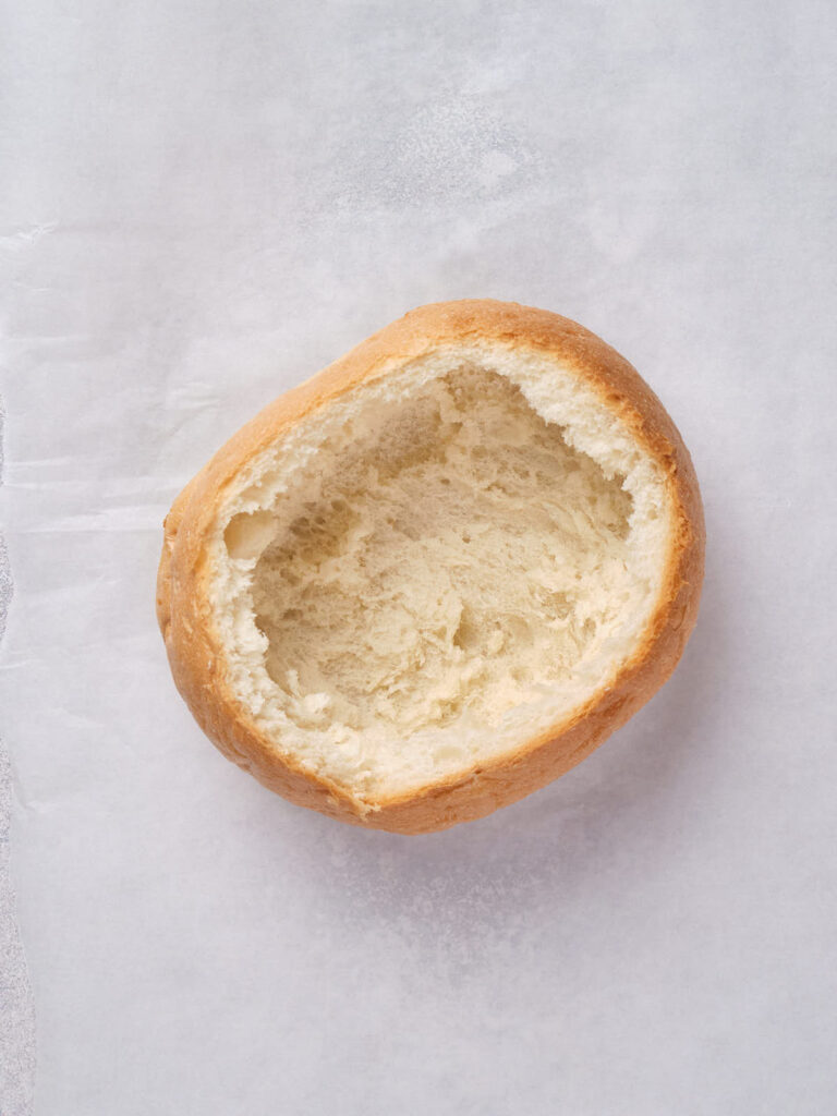 A hollowed round bread roll sits on a white surface, showing interior crumbs and a crispy crust.