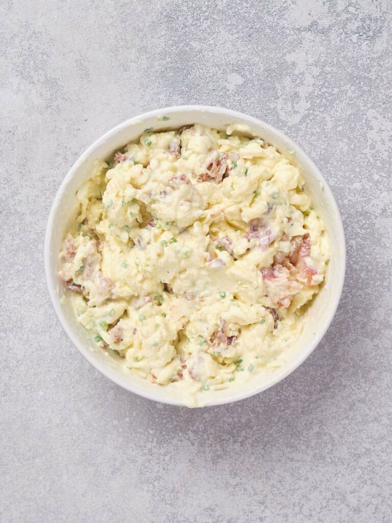 A bowl of creamy potato salad with chives and bacon on a gray surface.