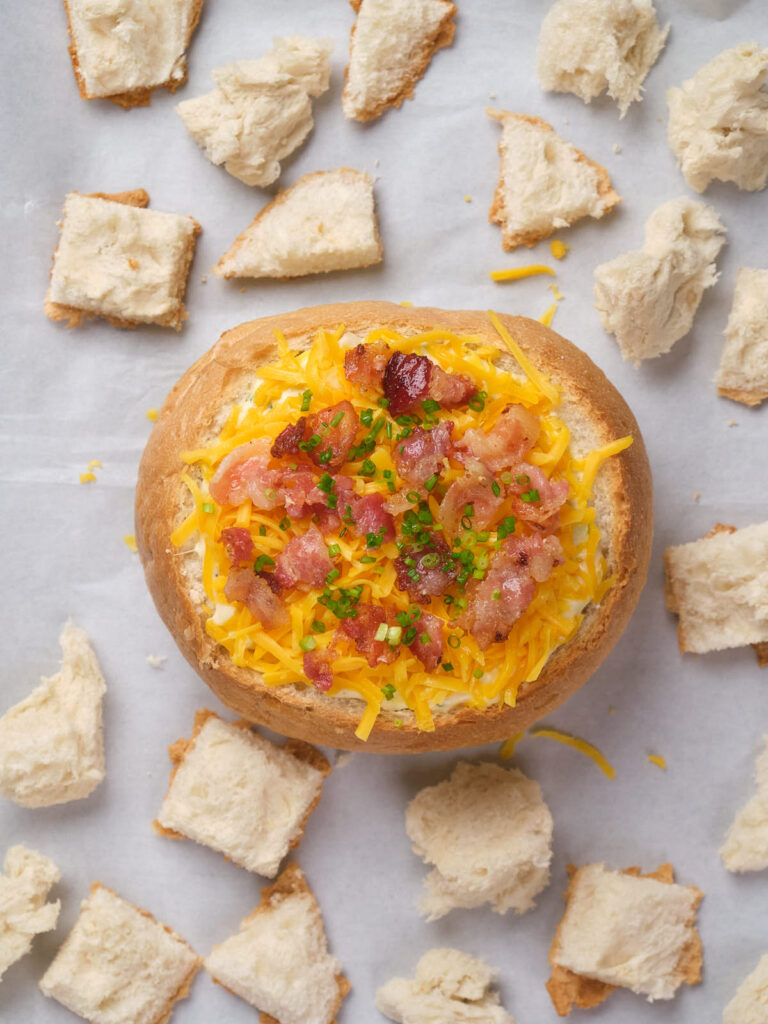 A bread bowl filled with soup, topped with shredded cheese, bacon, and chives.