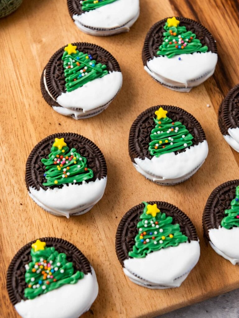 Chocolate sandwich cookies decorated with green frosting Christmas trees, white icing, colorful sprinkles, and yellow star toppers.