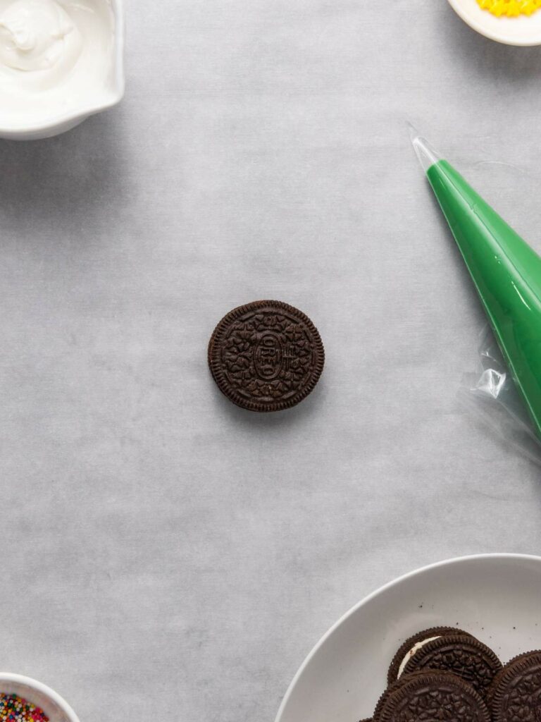 A single chocolate sandwich cookie on a gray surface surrounded by icing, sprinkles, and other ingredients.