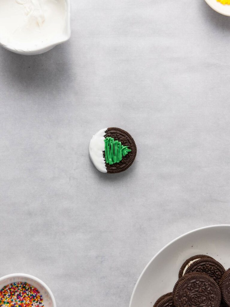 A chocolate sandwich cookie partially dipped in white icing, decorated with green icing in the shape of a Christmas tree, .