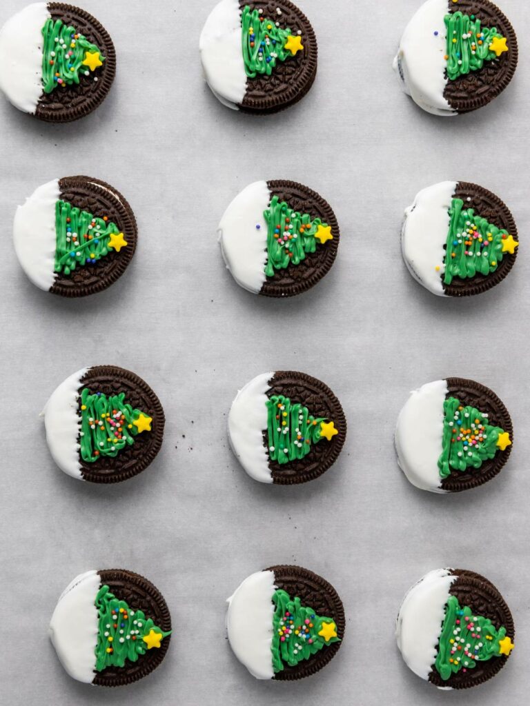 Twelve chocolate sandwich cookies half-dipped in white icing, each decorated with a green icing Christmas tree topped with a yellow star.