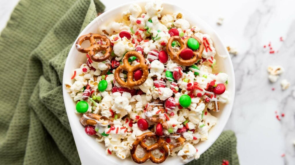 A bowl of popcorn mixed with pretzels, red and green candy pieces, white chocolate drizzle, and red sprinkles sits on a green textured cloth.