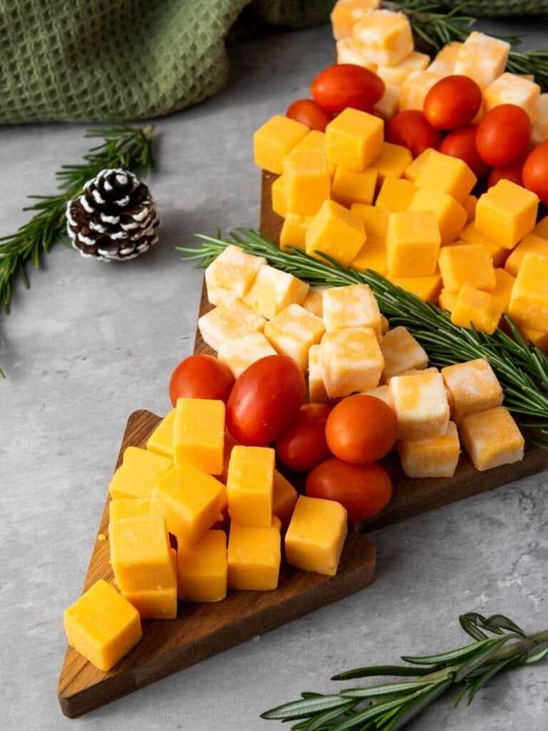 Cheese cubes, cherry tomatoes, and sprigs of rosemary arranged on a board, with a pinecone in the background.