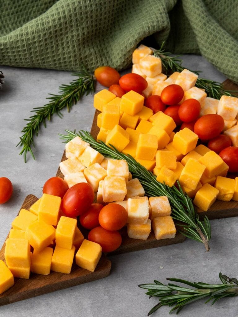 Cheese cubes and cherry tomatoes arranged on a wooden board with sprigs of rosemary, set on a gray surface.