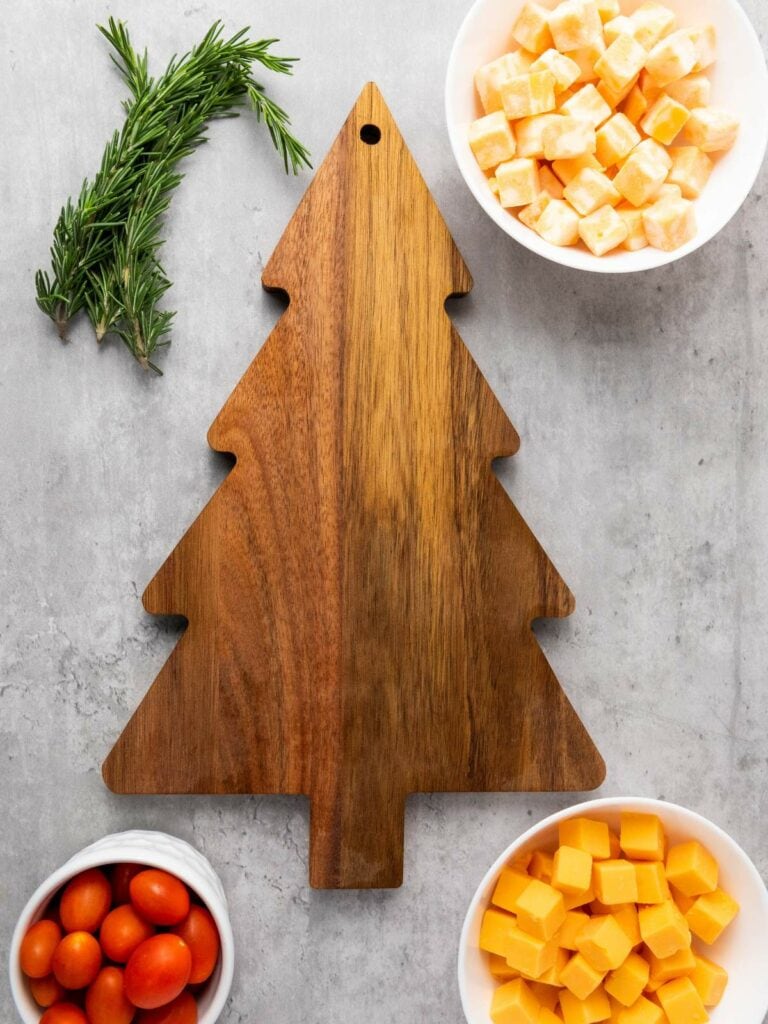 A wooden tree-shaped cutting board surrounded by bowls of cubed cheese, grape tomatoes, and rosemary on a gray surface.