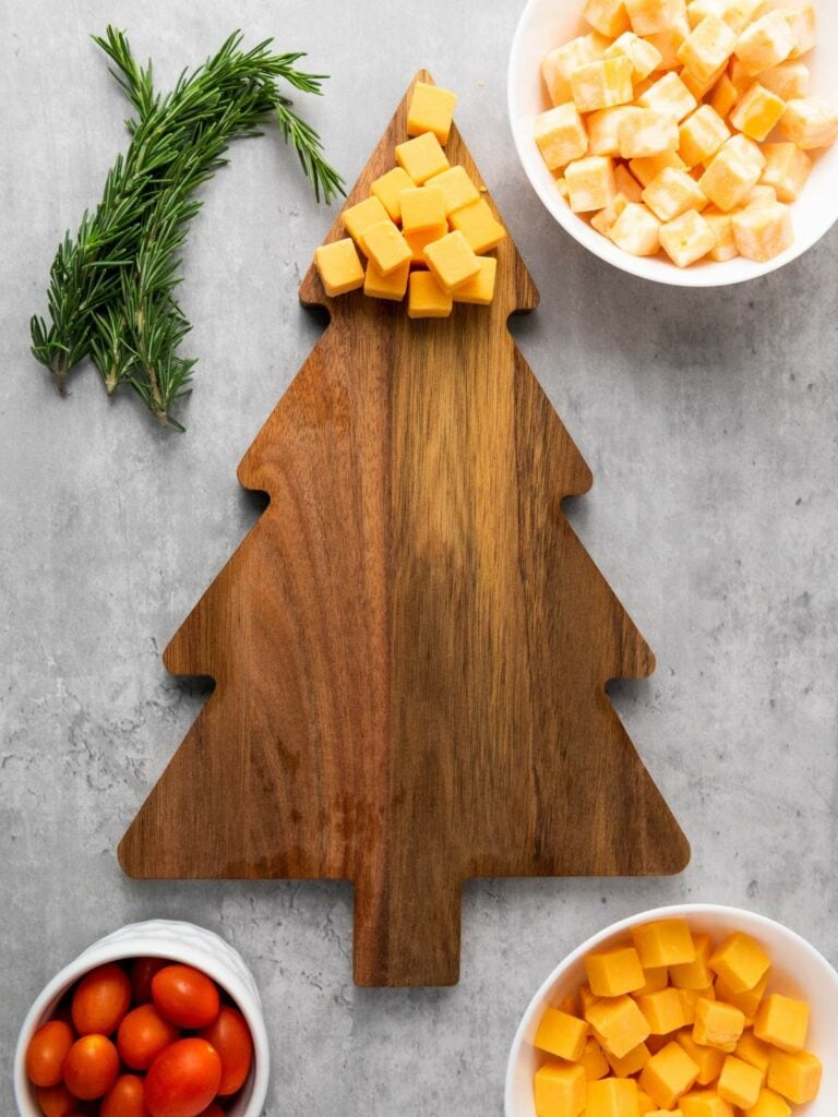 Wooden tree-shaped board with cubed cheese on top, surrounded by bowls of cheese cubes, tomatoes, and a sprig of rosemary.