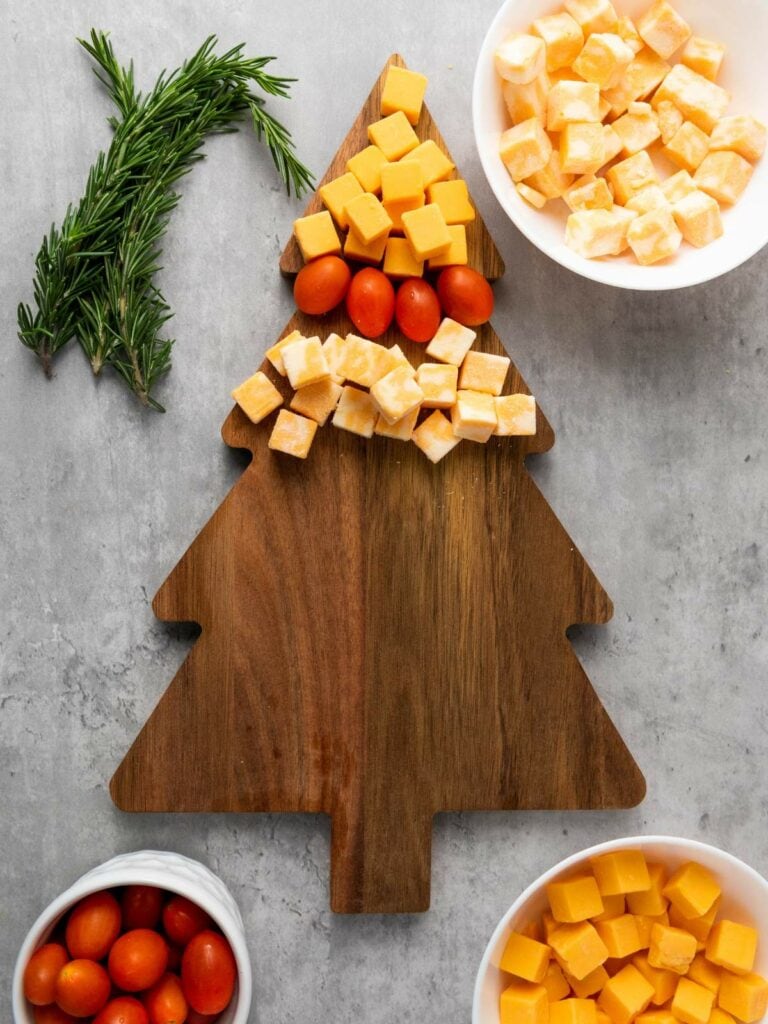 Cheese cubes and cherry tomatoes arranged on a tree-shaped wooden board, with sprigs of rosemary on the side.