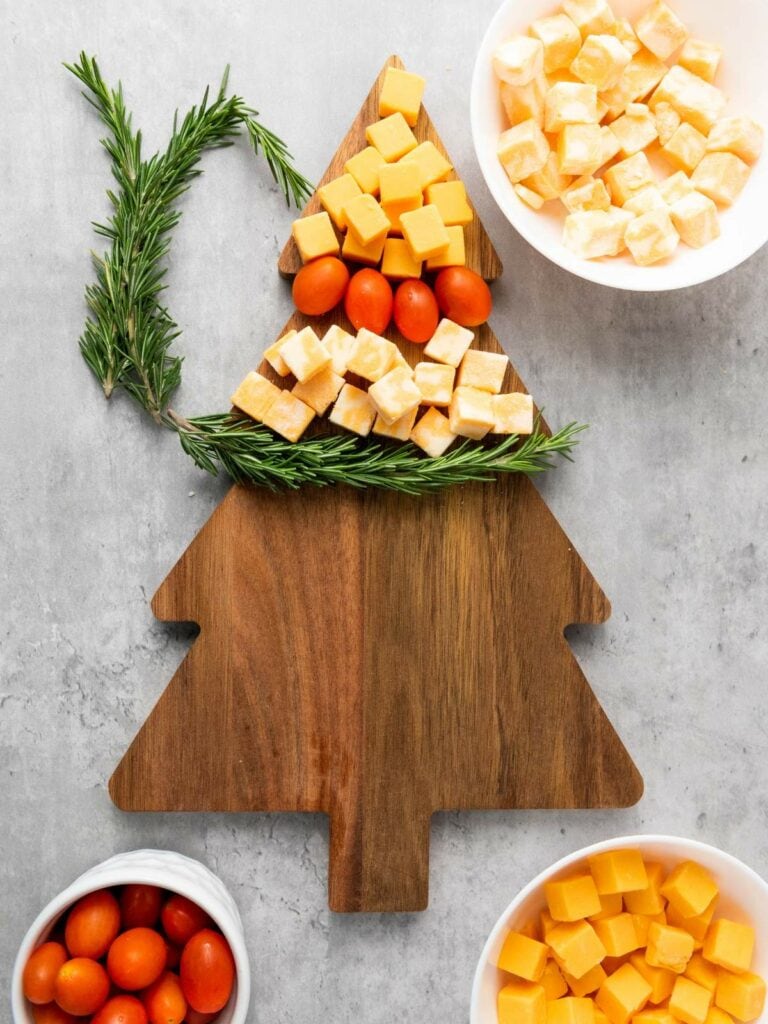 A tree-shaped wooden board with cheddar cubes, cherry tomatoes, and rosemary arranged on it.