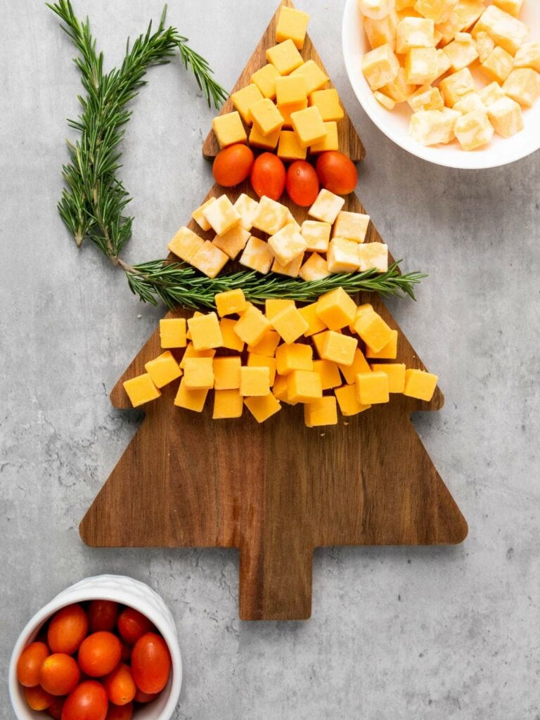 Cubed cheese and cherry tomatoes are arranged on a wooden tree-shaped board with rosemary on a gray surface.