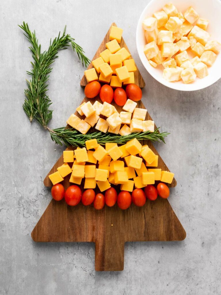 Wooden board with cheese cubes, cherry tomatoes, and rosemary arranged in a Christmas tree shape.