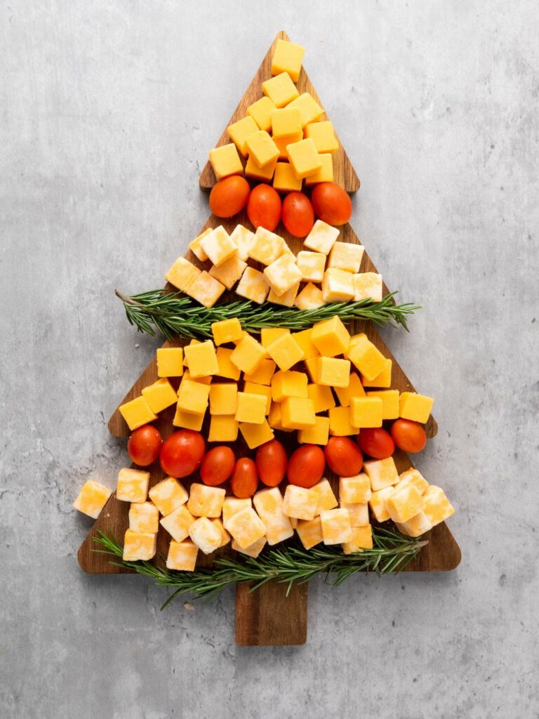 Cheese cubes and cherry tomatoes arranged in a Christmas tree shape on a wooden board, with rosemary sprigs as decoration.