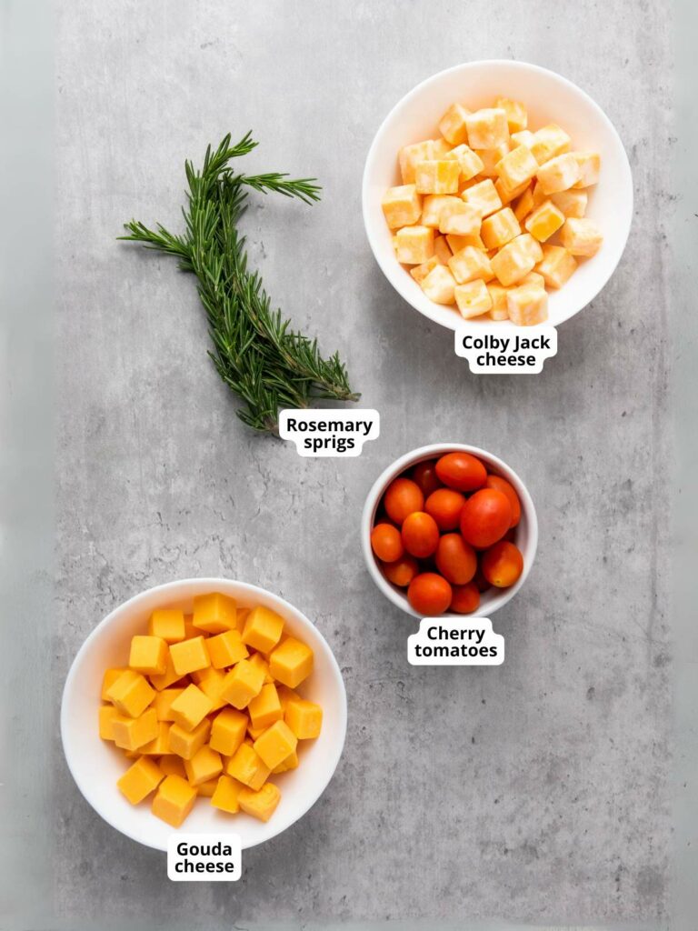 Three bowls with Colby Jack cheese, Gouda cheese, and cherry tomatoes on a gray surface, accompanied by sprigs of rosemary.