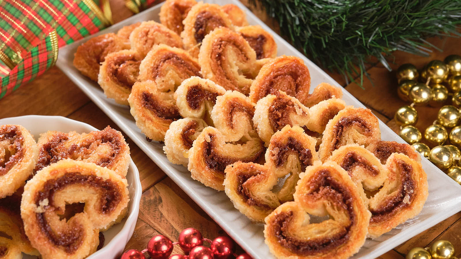 A rectangular platter with cinnamon palmiers and holiday decorations, including garlands and a gift wrap, is displayed.