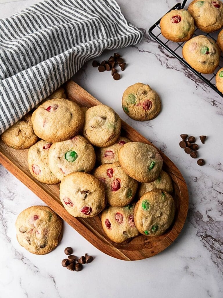 A wooden tray with assorted cookies, some with colorful candy pieces, sits on a marble surface.