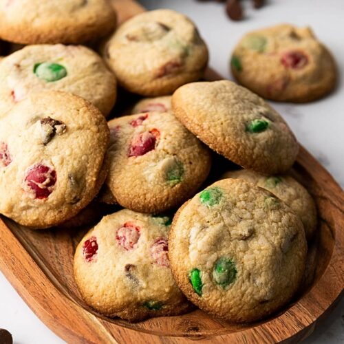 A wooden tray with freshly baked cookies containing red and green candies, placed on a marble surface.