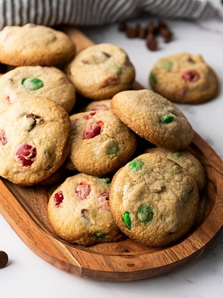 A wooden tray with freshly baked cookies containing red and green candies, placed on a marble surface.