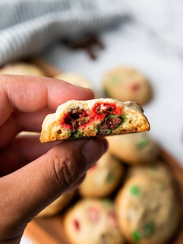 A hand holding a bitten cookie with red and green candy pieces, and more cookies in the background.