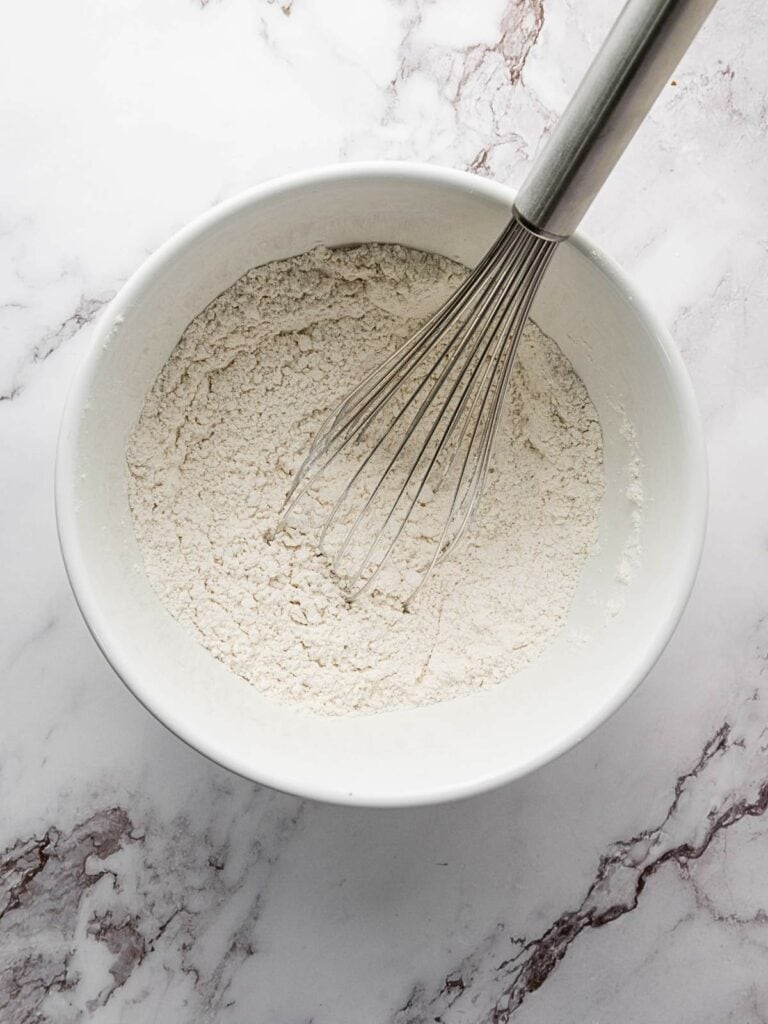 A whisk in a white bowl containing flour on a marble countertop.