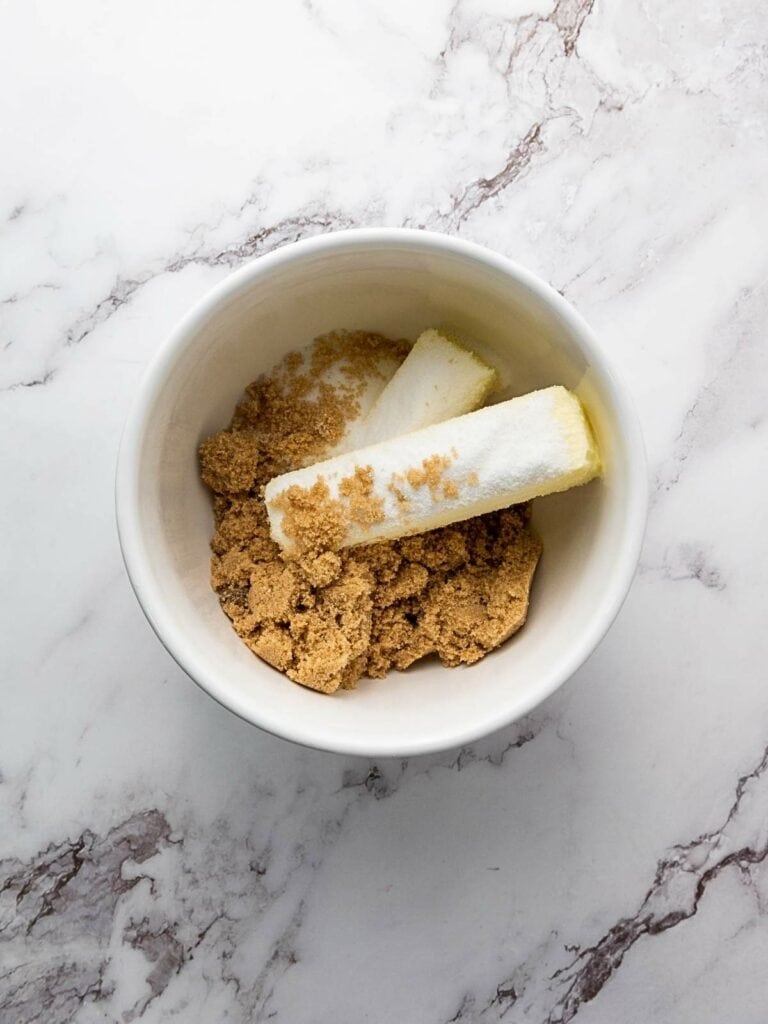 A bowl with brown sugar and two sticks of butter on a marble surface.