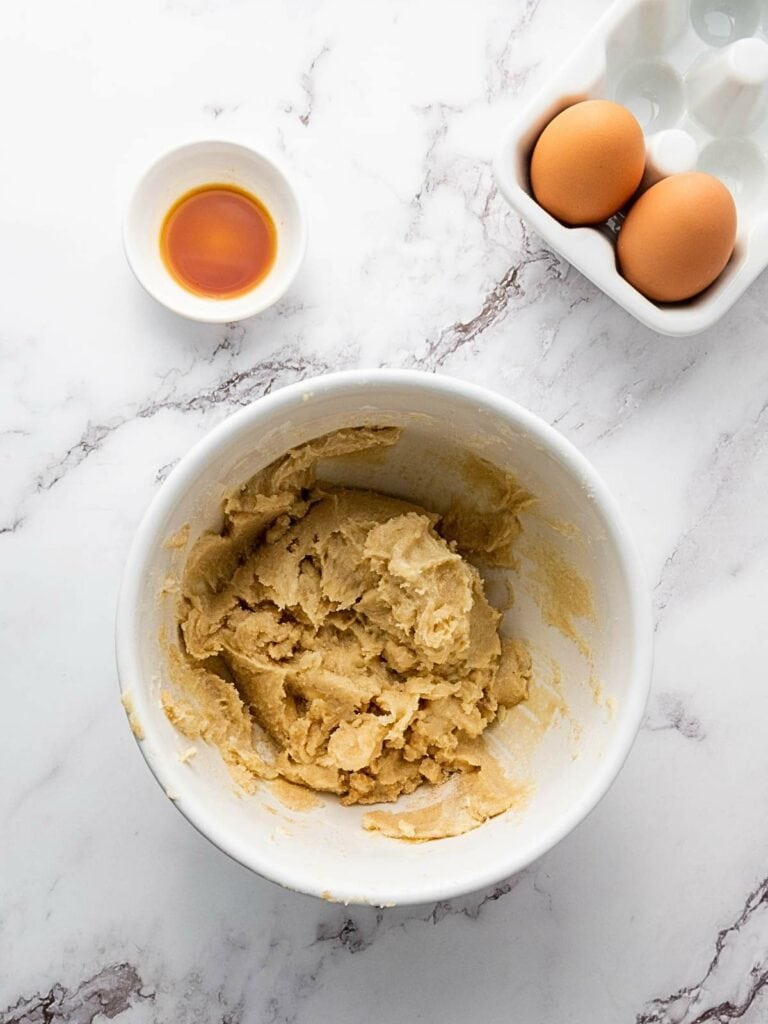 Bowl of cookie dough on a marble countertop, with two eggs in a carton and a small bowl of vanilla extract nearby.