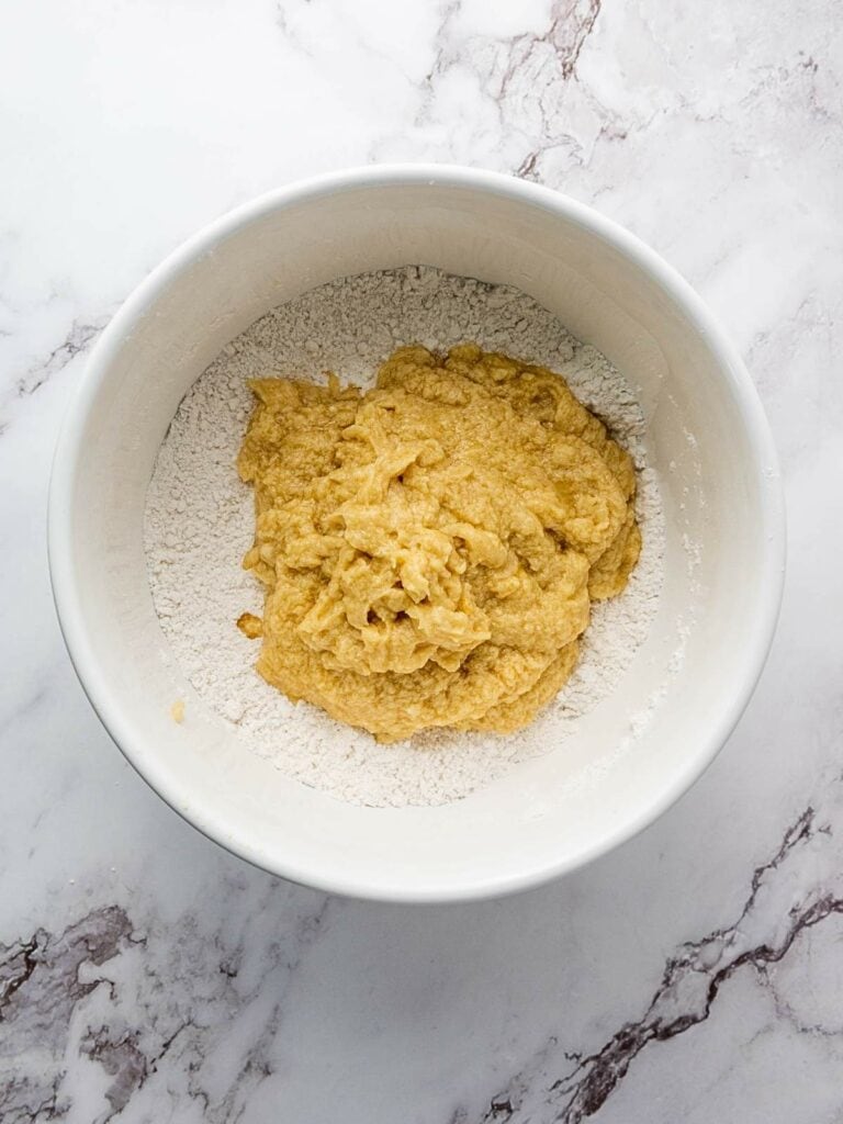 A white bowl contains dough on top of flour, placed on a marble surface.