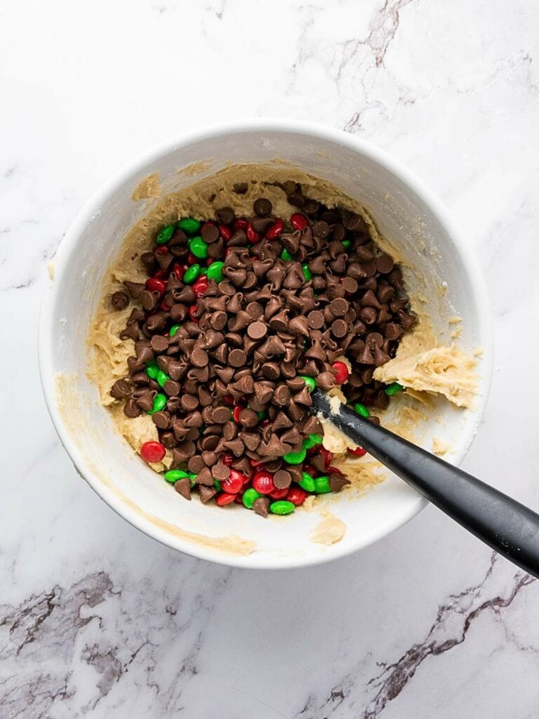 Bowl with cookie dough mixed with chocolate chips and red and green candies, on a marble surface.