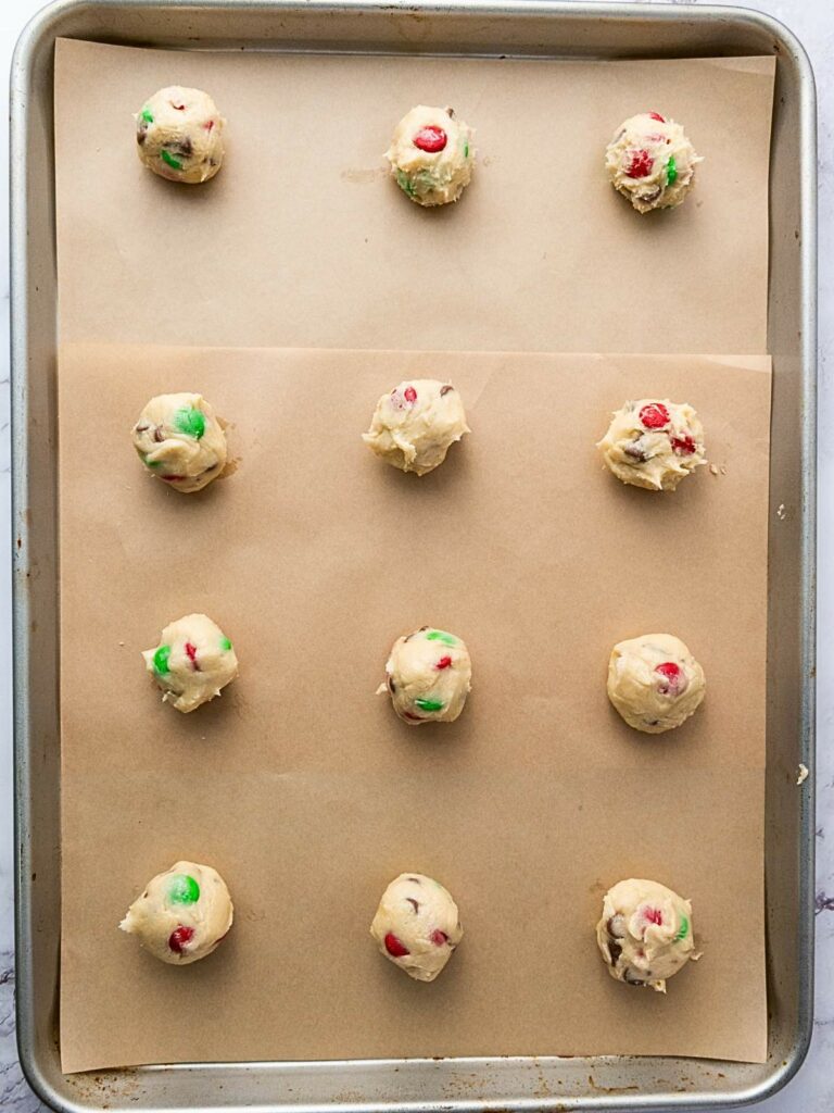 A baking tray with twelve evenly spaced scoops of cookie dough, some containing green and red candies, on parchment paper.